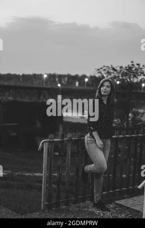 Immagine verticale in scala di grigi di una giovane donna che si trova vicino a un cancello di metallo in serata Foto Stock