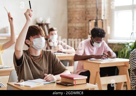 Gruppo di studenti in maschere protettive seduti alla deska e alzando le mani durante la lezione a scuola Foto Stock