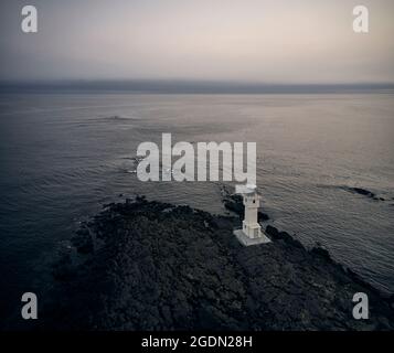 Vista sul drone del faro bianco situato sulla costa rocciosa vicino al mare ondeggiante al mattino tempestoso in Islanda Foto Stock