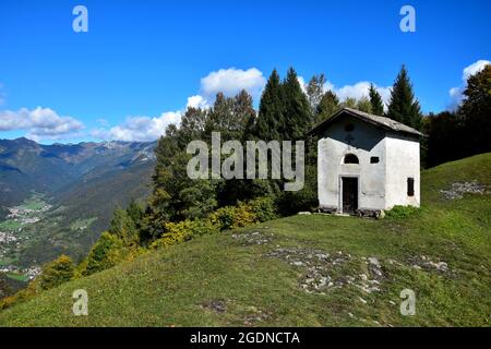 La piccola cappella San Martino sul Monte Corno in una bella giornata autunnale limpida. A sinistra alcuni piccoli centri abitati in una valle. Trentino, Italia. Foto Stock