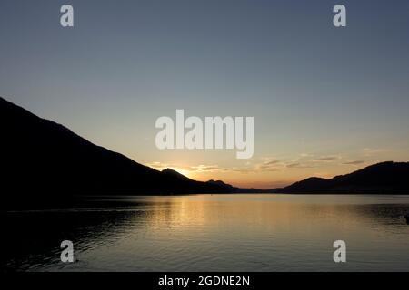 Pittoresco tramonto di fine estate. Basse nuvole gialle, ondate d'acqua di colore arancione e riflessi del lago trascurati dalla catena montuosa Alpina austriaca. Foto Stock