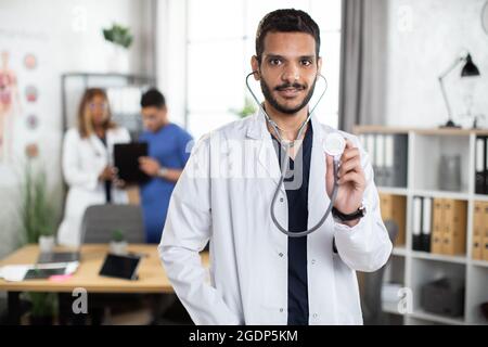 Primo piano inquadratura orizzontale di medico maschile medio orientale professionista in abito bianco, dimostrando il suo stetoscopio a macchina fotografica con sorriso. Due medici discutono la diagnosi in background Foto Stock