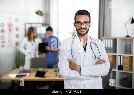 Scatto orizzontale con spazio per la copia. Concetto di medicina dove giovane allegro medico arabo soddisfatto uomo in piedi con le sue braccia incrociate in ufficio leggero con due medici multirazziali che parlano dietro Foto Stock