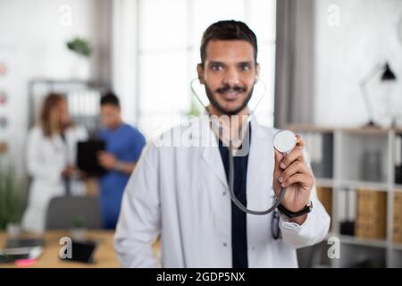 Primo piano visione offuscata del bel medico indiano maschile, in piedi con stetoscopio nella moderna sala d'imbarco dell'ospedale leggero con due diversi colleghi che parlano sullo sfondo. Concentrarsi sullo stetoscopio Foto Stock