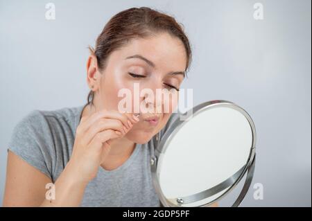 La donna caucasica guarda nello specchio e rimuove i baffi con l'aiuto di pinzette. Irsutismo. Foto Stock