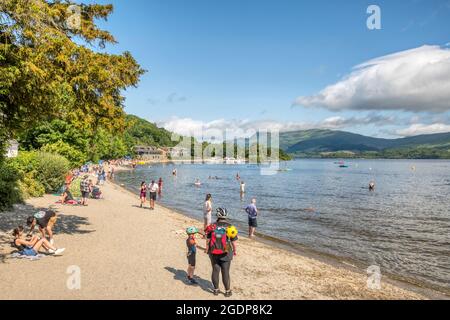 Persone in spiaggia a Luss accanto al Loch Lomond. Con Lodge su Loch Lomond e ben Lomond in background. Foto Stock