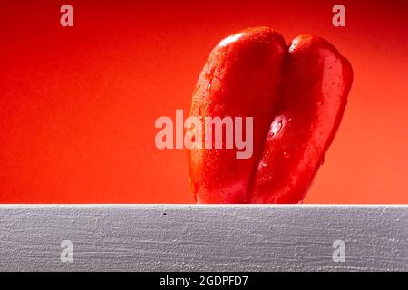 Fotografia di un peperone rosso bagnato su una tavola bianca e uno sfondo rosso. La foto viene scattata in formato orizzontale. Foto Stock