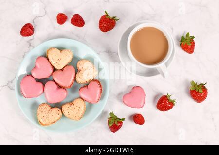 I dolci francesi di macaron a forma di cuore rosa e beige accanto alla tazza da caffè e alla frutta di fragole e lamponi Foto Stock