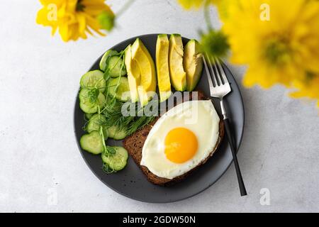 Deliziosa colazione con avocado, cetrioli freschi e micrograni di piselli con aneto, uova fritte su pane tostato a grani neri, vista superiore dei piatti con cibo Foto Stock