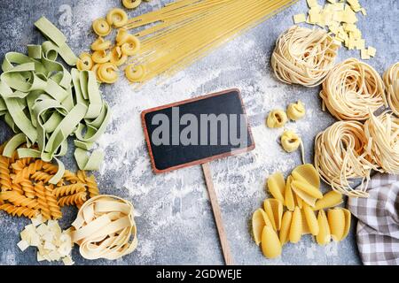 Etichetta in lavagna bianca tra tipi misti e forme di pasta italiana su pietra grigia, sfondo. Spazio di copia. Foto Stock