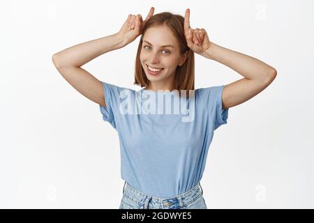 Immagine di sciocca e carina giovane donna che si diverte, mostrando corna toro gesto sulla testa e sorridendo felice, essendo giocoso, in piedi in t-shirt contro bianco Foto Stock