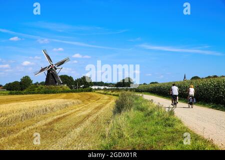 Beesel, Paesi Bassi - 9 luglio. 2021: Vista sulla pista ciclabile con ciclisti nel paesaggio rurale olandese maas con mulino a vento (birra Molen de grauwe) contro blu Foto Stock