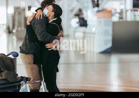 Donna in maschera di faccia che abbruta l'uomo al cancello d'arrivo dell'aeroporto. Donna che abbraccia e accoglie l'uomo dopo la pandemia in aeroporto. Foto Stock