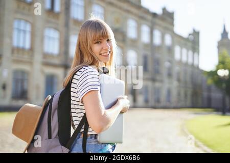 Studio all'estero, studio di scuola superiore o concetto educativo. Carino felice smiling blonde college o studentessa universitaria con zaino, cappello e laptop nel campus in estate. Immagine di alta qualità Foto Stock