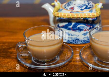 Tè fatto con latte servito in una tazza di vetro e piatto su un tavolo di legno da una teiera con manico in background Foto Stock