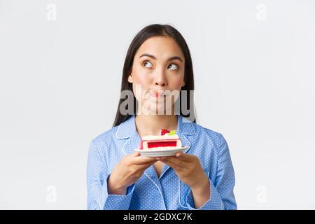 Primo piano di bella ragazza asiatica pensierosa in piedi di notte in pajama con piatto di torta, guardando in alto angolo sinistro decidere, cant resistere tentazione Foto Stock