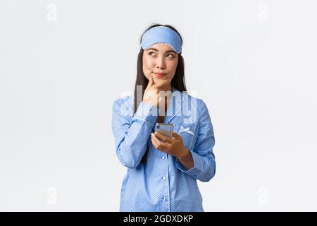 Tecnologia, persone e concetto di svago domestico. Pensieroso e creativo cute blogger femminile, ragazza asiatica in pigiama e la maschera da sonno hanno idea, pensando Foto Stock