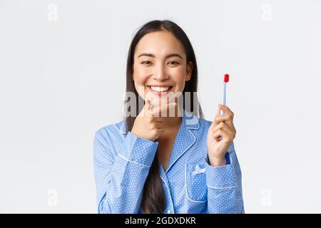 Primo piano di attraente donna asiatica in pajama blu pensando mentre spazzolando i denti con spazzolino da denti, sorridendo e toccando mento ponderare grande idea Foto Stock