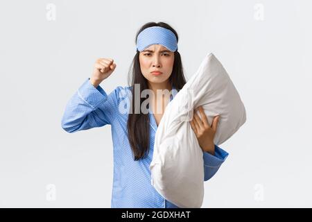 Arrabbiato e spossato ragazza asiatica spossata lamentando forti vicini al dormitorio, indossando la maschera da sonno e pajama, tenere cuscino e minacciare qualcuno Foto Stock