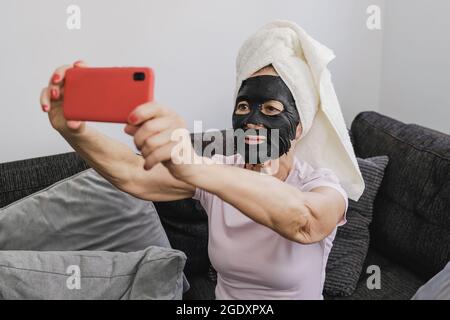 Donna anziana che usa la maschera facciale di carbone mentre prende un selfie con il telefono mobile indoor a casa - cura della pelle concetto sano di stile di vita - fuoco sul viso Foto Stock