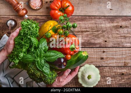 Mano femminile che tiene un cesto con verdure fresche e frutta su vecchio sfondo rustico in legno, vista dall'alto. Spazio per il testo. Cibo cottura sfondo e m Foto Stock