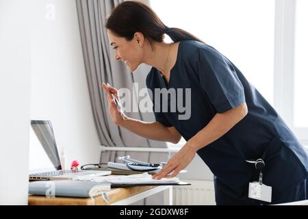 Una ragazza sorridente dottore in piedi al tavolo e mostrando il suo palmo alla macchina fotografica portatile nella stanza di fronte alla finestra Foto Stock
