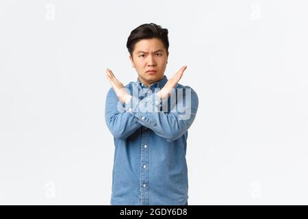 Serio-guardando interessato studente asiatico di sesso maschile mostrando gesto proibito, facendo croce segno di fermare qualcuno, in disaccordo e vietare l'azione, dicendo di no Foto Stock
