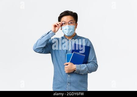 Covid-19, prevenire i virus, e la distanza sociale al concetto universitario. Entusiasta uomo asiatico felice in maschera medica in piedi con taccuini e. Foto Stock