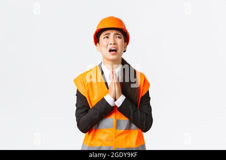 Disperato e sconsiderata responsabile della costruzione femminile asiatica, architetto che supplicava dio, indossando casco di sicurezza, tenendo le mani in preghiera, supplicando, chiedendo Foto Stock