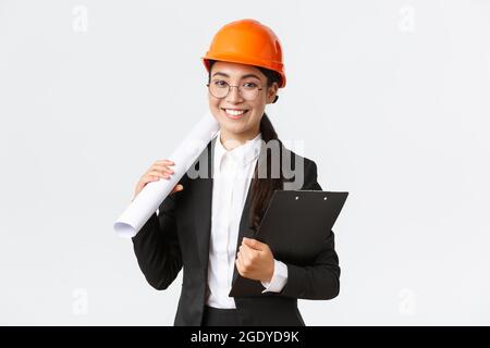 Architetto femminile asiatico di successo in tuta da lavoro e casco tenendo blueprint e appunti con note, ispettore guardando lavori di costruzione Foto Stock