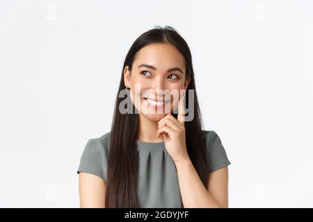 Primo piano di creatività e sognante sorridente bella donna asiatica pensare, l'imaging qualcosa di interessante in mente, guardando a sinistra e toccante guancia, meditare Foto Stock