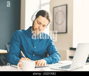 Lavoratore maschile in ufficio vestito con abiti casual in cuffie guardando online webinar su laptop Foto Stock