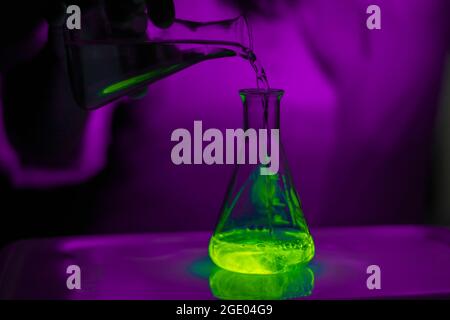 Uno scienziato che lavora con una soluzione fluorescente verde in un matraccio conico di vetro in un laboratorio biomedico scuro per lo sviluppo dell'assistenza sanitaria Foto Stock