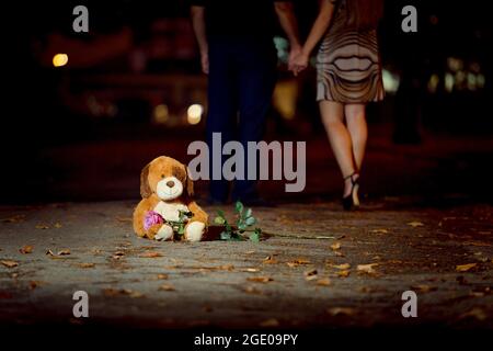 Un orsacchiotto si siede sull'asfalto e tiene una rosa fresca. Sullo sfondo delle gambe di una coppia amorevole di uomo e donna. Foto Stock