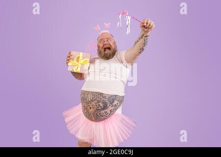 L'uomo eccitato con il peso eccessivo in costume fata tiene il bastone magico e presente su sfondo viola Foto Stock