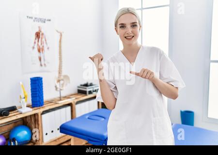 Giovane donna caucasica che lavora in clinica di recupero del dolore puntando alla schiena dietro con la mano e i pollici in su, sorridente sicuro Foto Stock