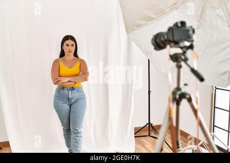 Giovane bella donna ispanica che si pone come modello in studio di fotografia scettica e nervosa, disapprovando l'espressione sul viso con le braccia incrociate. Negativo Foto Stock