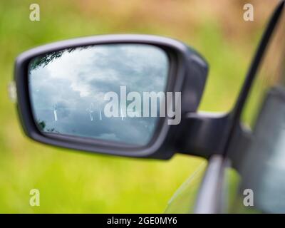 Specchietto retrovisore sinistro della vettura con vista poco chiara sul sedile del conducente. La strada dell'entroterra con alberi verdi sfocati Foto Stock