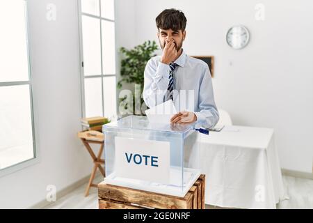 Uomo ispanico con la barba votando mettere inviluppo in una scatola di voto odore qualcosa di stinoso e disgustoso, odore intollerabile, respiro trattenuto con le dita Foto Stock