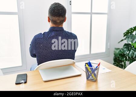 Giovane uomo ispanico con barba che lavora in ufficio con un computer portatile in piedi indietro guardando via con le braccia incrociate Foto Stock