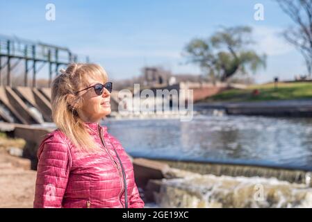 Vista di un'allegra donna adulta che guarda lontano con occhiali da sole in piedi nel parco con un laghetto dietro in una giornata di sole. Foto Stock