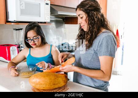 Mamma latina affettare frutta con la figlia adolescente con sindrome di Down in cucina, nel concetto di disabilità in America Latina Foto Stock