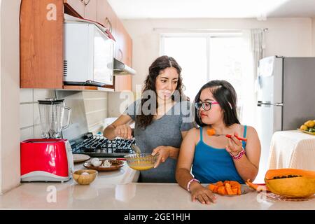 Teen girl messicana con sindrome di Down mangiare frutta e sua madre cucina a casa, in concetto di disabilità in America Latina Foto Stock