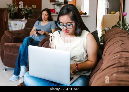 Ragazza latina adolescente con sindrome di Down studiare sul computer seduto sul divano con la mamma a casa, in concetto di disabilità in America Latina Foto Stock