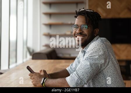 Sorridente giovane dipendente africano in ufficio siede sul posto di lavoro con uno smartphone Foto Stock