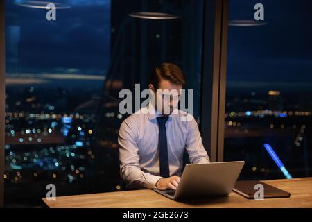 L'uomo d'affari si siede sul posto di lavoro fino a tardi utilizzando il computer portatile Foto Stock