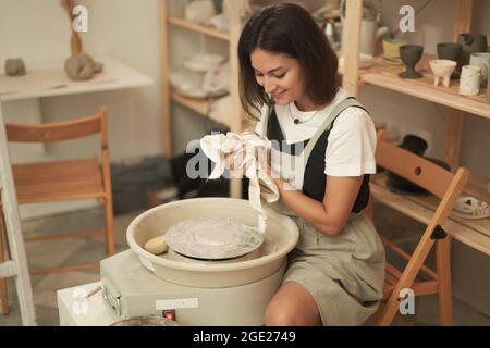 Vista laterale del contenuto giovane artista ceramica femminile strofinando le mani con un panno mentre si siede alla ruota in ceramica dopo il lavoro con argilla in studio Foto Stock
