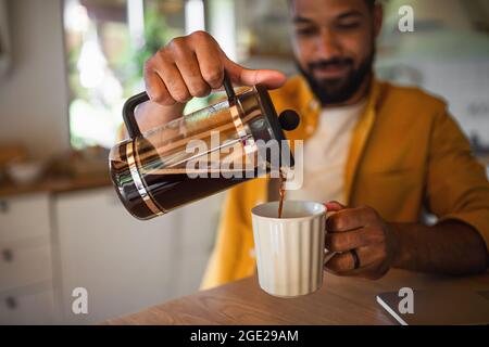 Giovane uomo che versa il caffè dalla pressa francese che lavora all'interno, concetto di ufficio domestico. Foto Stock