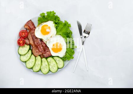 Uova di pollo fritte con pancetta e verdure su un tavolo grigio. Concetto di colazione. Vista dall'alto. Cibo di sottofondo al mattino. Cetrioli e toma freschi a fette Foto Stock