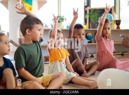 Gruppo di bambini piccoli della scuola materna seduti al piano interno in classe, alzando le mani. Foto Stock